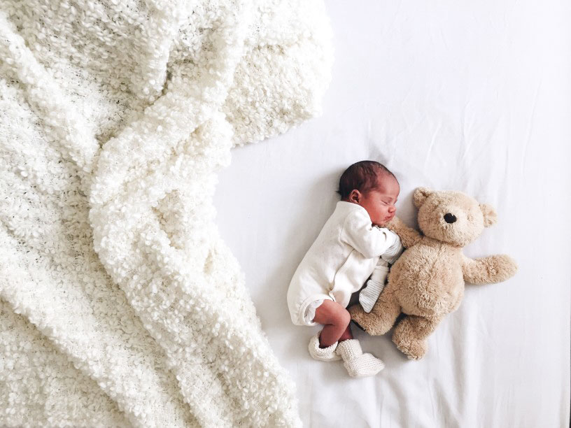 Newborn infant sleeping next to a teddy bear and a plus blanket in neutral colors