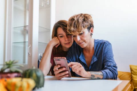 Mother and daughter sit at the kitchen table and look at a mobile phone.
