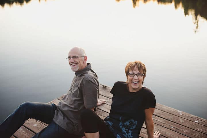 Middle-aged, retired couple sitting together laughing outside on a dock over a lake