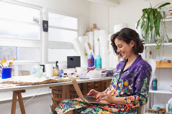 Young female artist smiles as she works on her laptop in her painters' studio