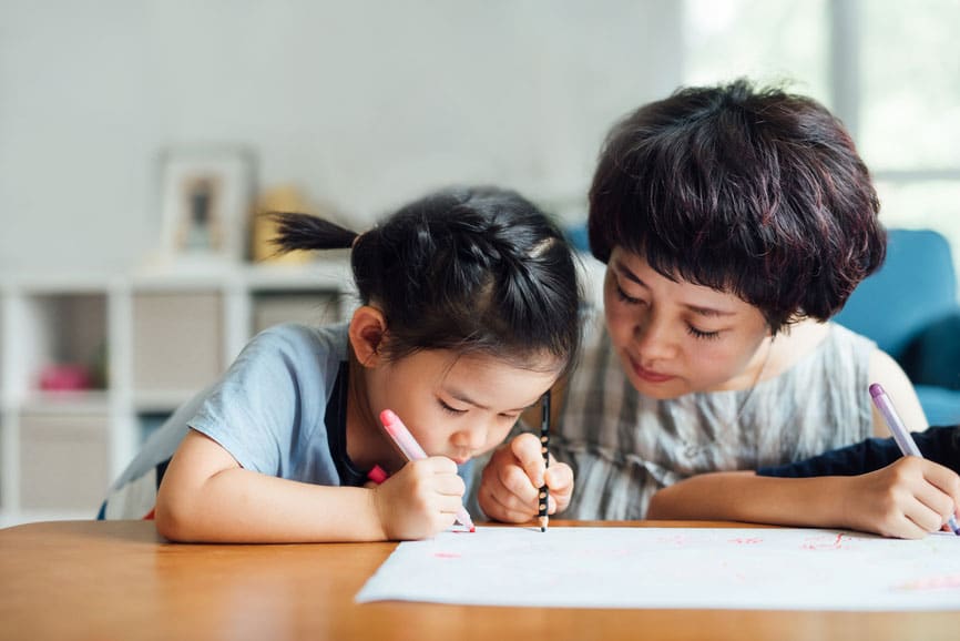 An Asian woman sitting at a table coloring with her toddler daughter