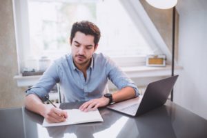 A man works on his finances, making notes on paper while referencing his laptop