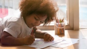 Kindergartener sits next to a window, coloring with colored pencils