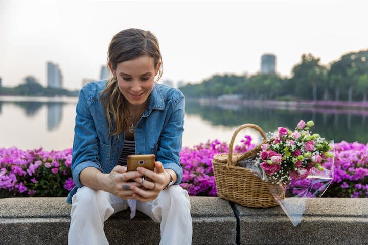 A woman sits, using her phone, on a wall enclosing flowers, behind which is a lake enclosed by a high-rise and tree skyline