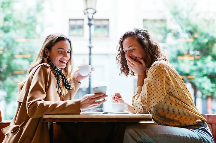 Two young ladies laugh over something on their phone while they have coffee in a cute shop