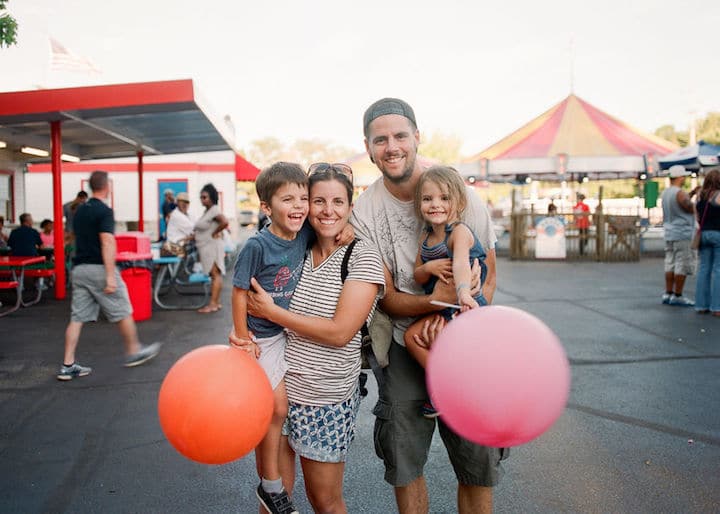 Parents pose with their two children at an amusement park, parents holding the kids and the kids holding oversized balloons