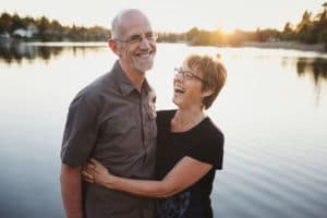 An older, retired couple hold their arms around each other, grinning and laughing, as they stand by a lake