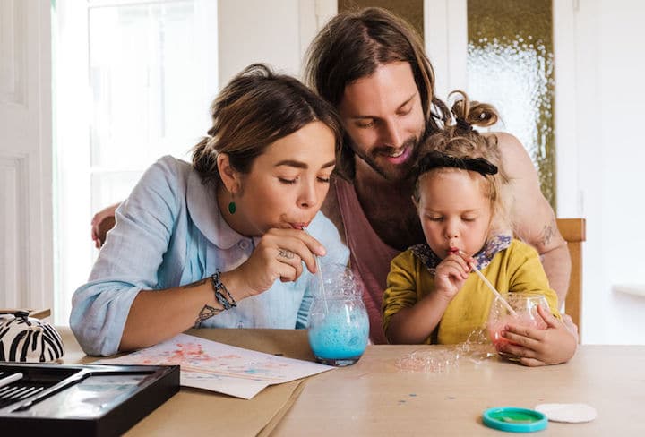 Young parents and little girl blowing into straws and making colorful foam in glasses with soap bubbles liquid