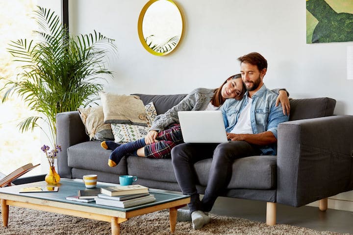 Young woman curls up on couch, leaning on her partner's shoulder while he works on his laptop