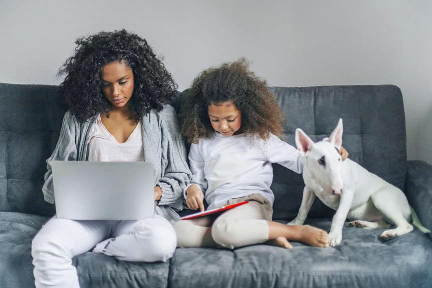 Anne dizüstü bilgisayarda ve kızı tablette kanepede bir köpekle oturuyor.