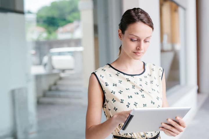 Young businesswoman works outside her office on a tablet, dark hair pulled back and wearing a floral-print sleeveless dress