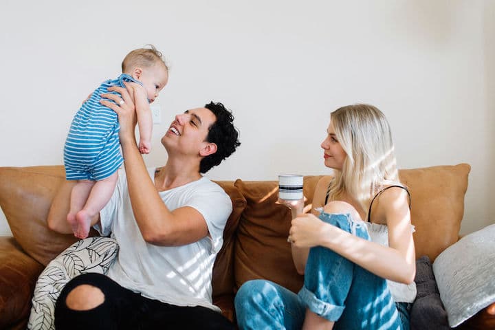 Dad lifts baby up into air as mom watches, sipping coffee