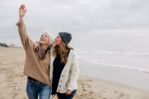 Adult daughter wraps her arm around her mom's shoulders while her mom reaches towards the sky and they both enjoy a winter walk on the beach