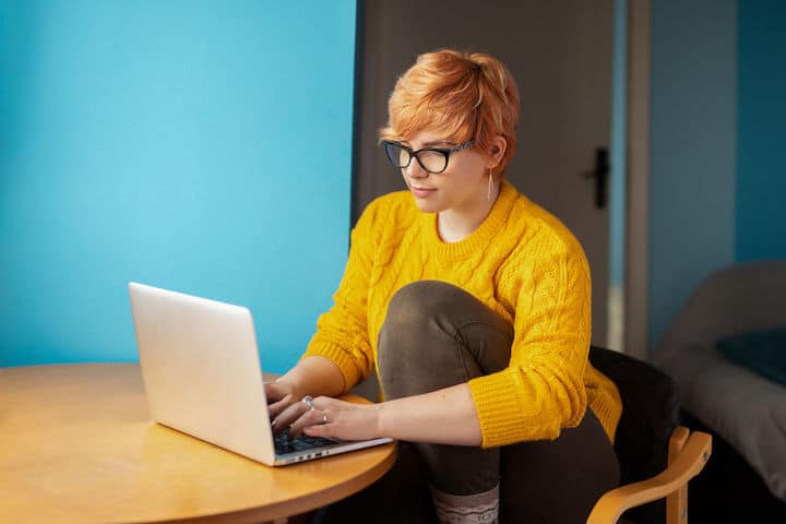 Young woman with pixie haircut and yellow sweater works from home on her laptop