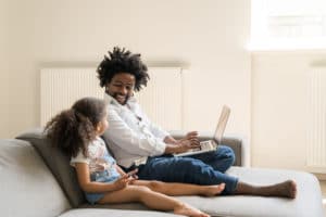 African American father sitting on his couch looking at his laptop while his daughter sits next to him