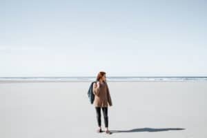 Woman sanding alone on a beach, looking out into the ocean while carrying her jacket.