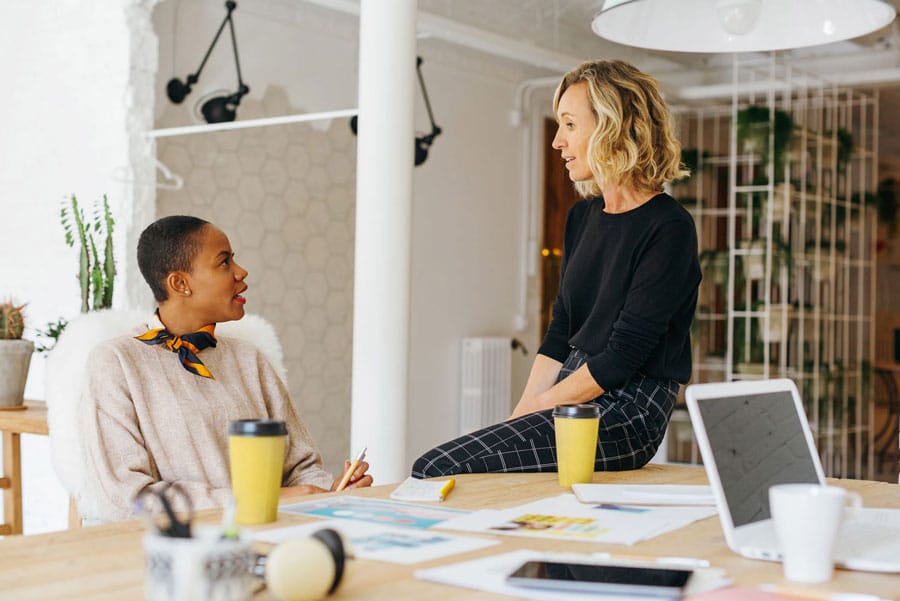 One white female coworker chatting with one black female coworker at her desk with coffees and laptops on the desk setting