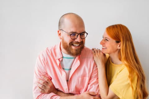 Portrait of married couple, handsome bald man and beautiful redhead woman, posing in front of white wall, looking at each other and smiling