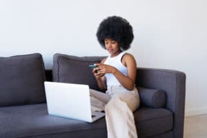 African American lady sitting on sofa and browsing netbook. Woman with Afro hairstyle using smartphone while working on remote project at home