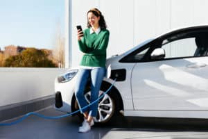 Full body young female smiling and browsing mobile phone while leaning on charging electric vehicle on sunny day on station