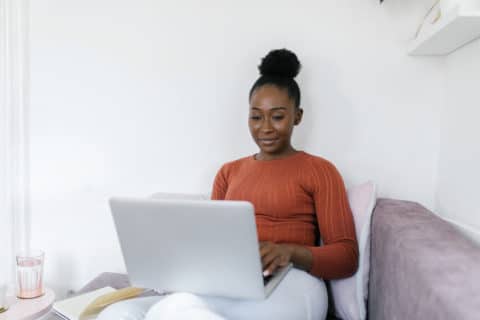 Beautiful African American woman working from home. She is sitting on the sofa in a cozy living room with laptop in her lap.