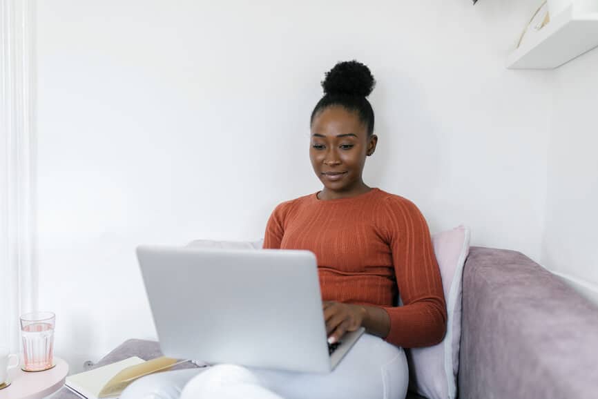 Beautiful African American woman working from home.  She is sitting on the sofa in a cozy living room with a laptop on her lap.