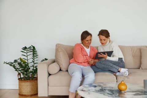 Young woman in casual clothes showing video on tablet to mature mother while resting on comfortable sofa at home together