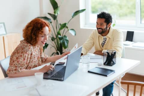Happy diverse man and woman with laptop and paper smiling and working on project together while sitting at table in bright workspace
