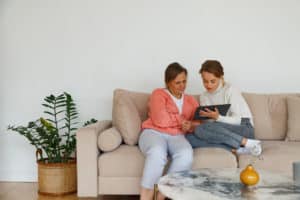 Young woman in casual clothes showing video on tablet to mature mother while resting on comfortable couch at home together