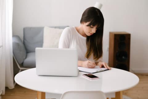 Jeune femme d'affaires assise à une table avec un ordinateur portable et un téléphone portable dessus.  Elle écrit sur le presse-papiers