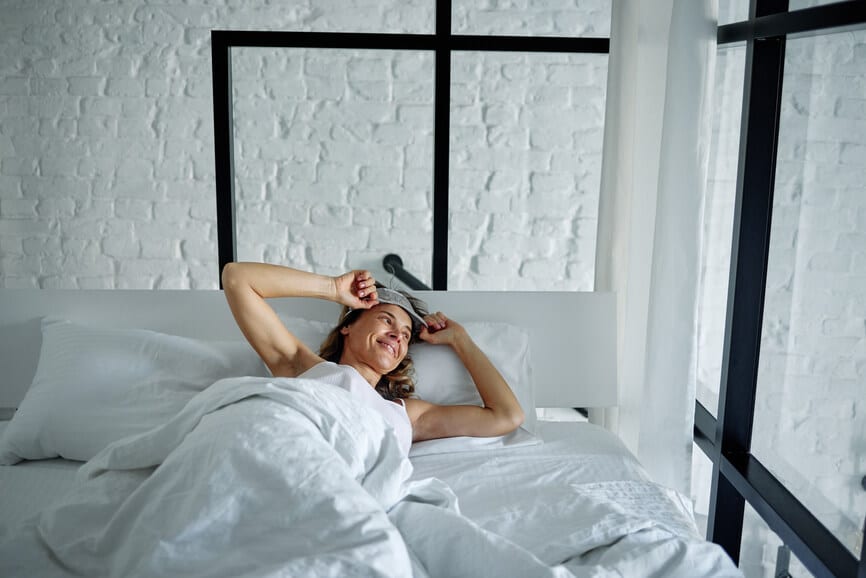 Front view of happy woman, hugging sleeping mask, reclining on bed in spacious room and looking out window in the morning
