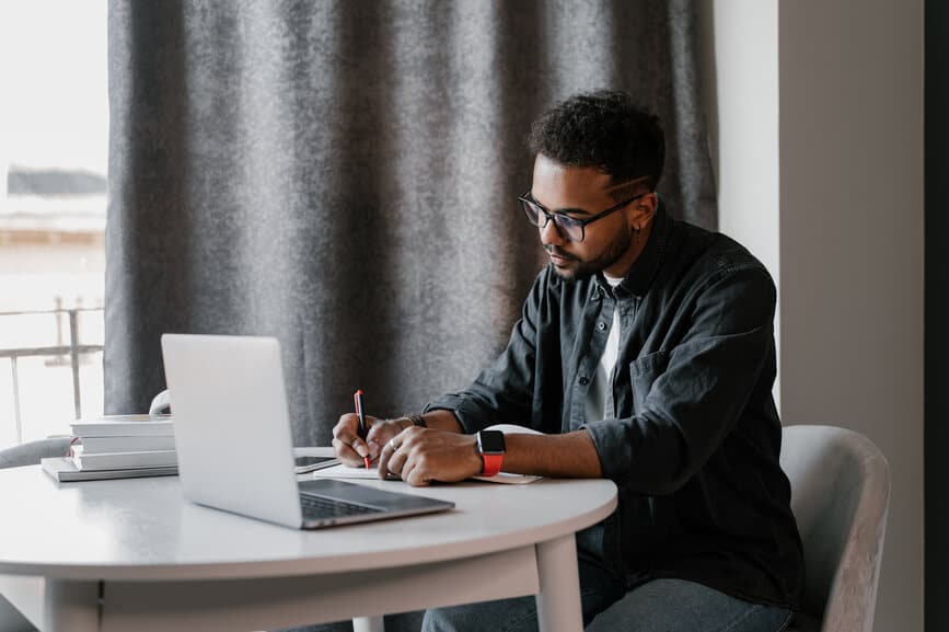 Black man with glasses sitting at table with laptop and writing in notebook while working on freelance project at home