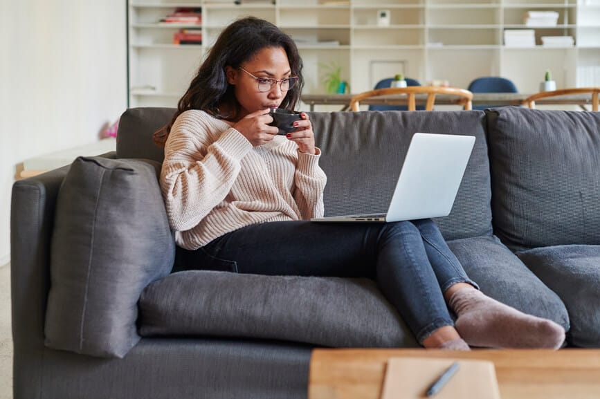 Genç Afro-Amerikan kadın bir dizüstü bilgisayarla internette çalışıyor ve evdeki oturma odasındaki kanepede otururken kahve içiyor.