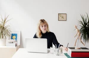 A portrait of a beautiful blonde haired woman working in a white airy office. She has a relaxed confident expression and is looking towards a piece of paper she is writing on. She is wearing a blue shirt polka dot retro shirt and is sitting behind her laptop at her desk.