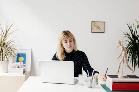 A portrait of a beautiful blonde haired woman working in a white and airy office.  She has a relaxed confident expression and looks towards a piece of paper she is writing on.  She is wearing a blue polka dot retro shirt and is sitting behind her laptop at her desk.