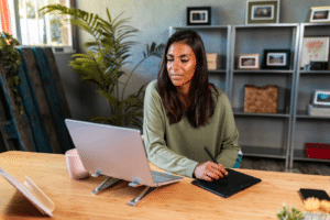 Self-employed hispanic woman using graphic tablet and netbook while working from home office on project online sitting at desk