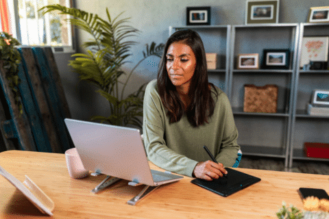 Self-employed hispanic woman using graphic tablet and netbook while working from home office on online project sitting at desk