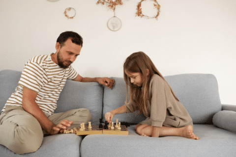 Bearded father watching smart kid move while sitting on sofa and teaching daughter to play chess on holidays at daytime at home.