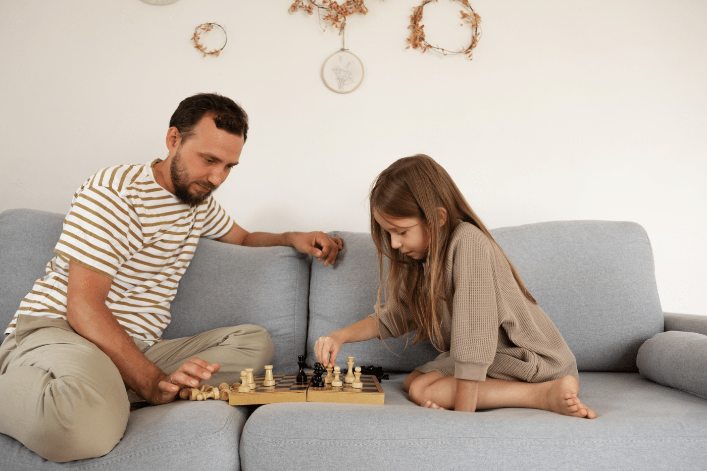 Bearded father watching smart kid making move while sitting on couch and teaching daughter to play chess on weekend day in daytime at home.