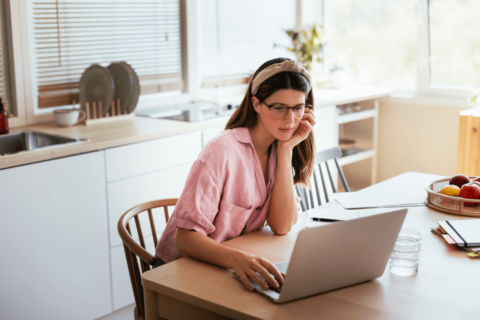 Young female freelancer in glasses leaning on hand and thinking while sitting and table and browsing netbook during work at home