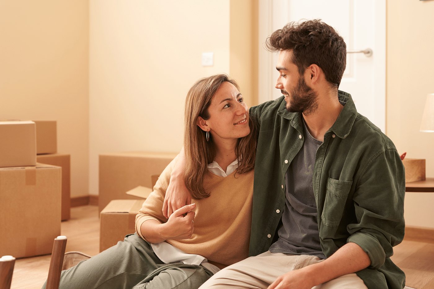 Young man hugging and looking at girlfriend sitting on floor near cardboard boxes in new apartment
