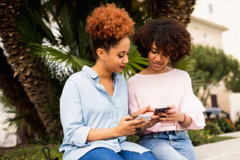 Two female friends using social media on smartphones and relaxing outdoors while sitting on a bench in the city. Leisure time outdoors.