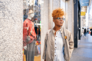 Modern Black Woman With Curly Hair And Glasses Doing Shopping Walking In Front Of The Showcase Of A Shop On The Street Of The City.