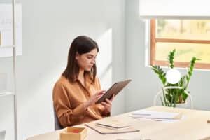Focused trendy lady in casual clothes sitting at table and using tablet while working on startup project in light home office
