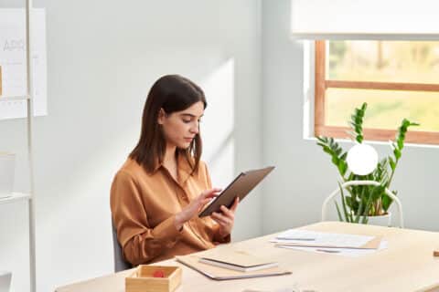 Focused trendy lady in casual clothes sitting at table and using tablet while working on startup project in light home office