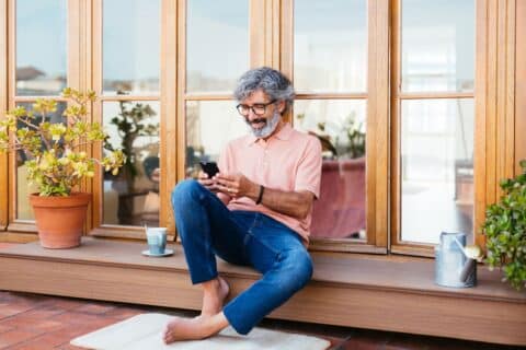 Senior man with gray beard wearing stylish salmon polo shirt and blue jeans barefoot using smartphone on cold city terrace