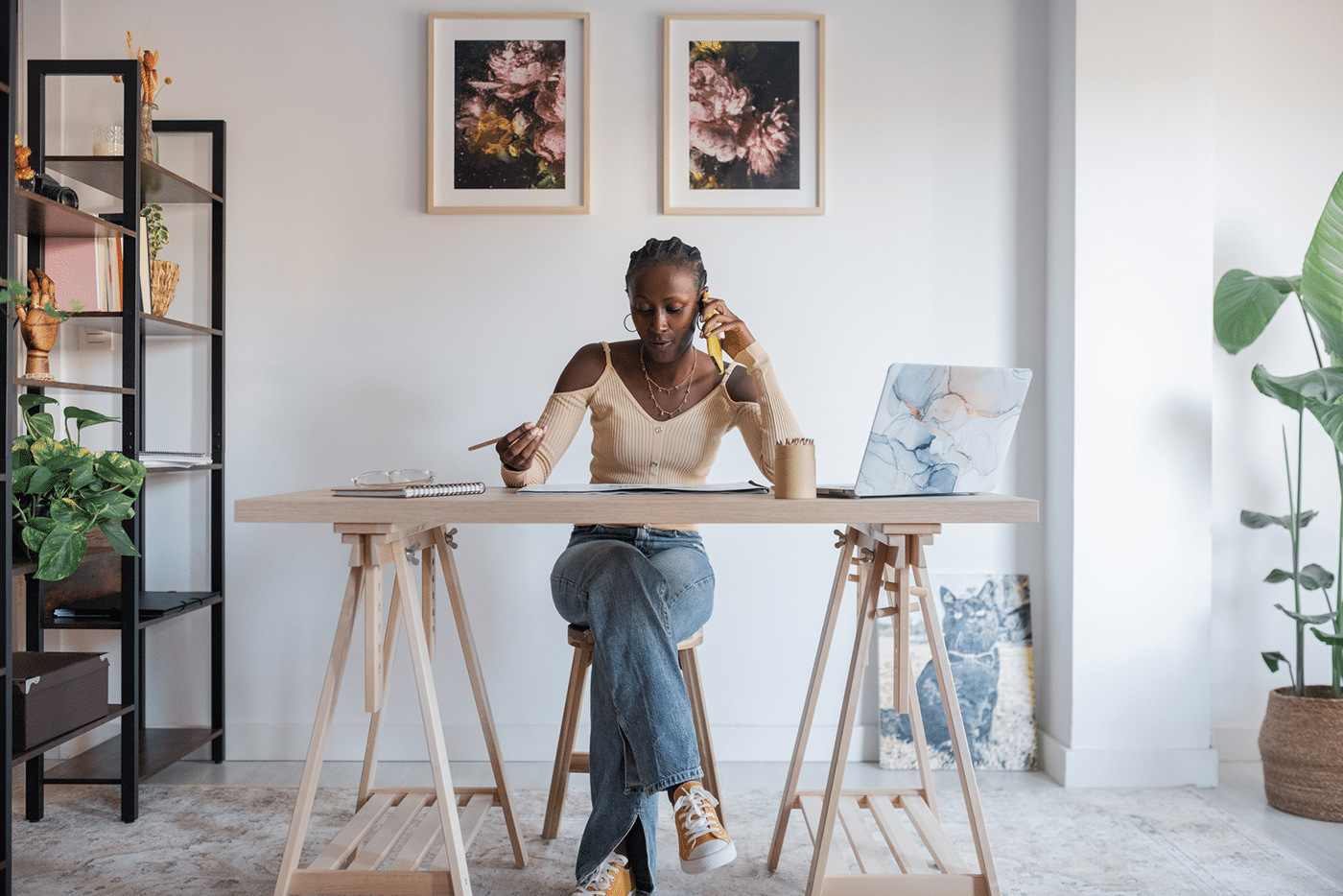African American woman in casual clothes examining blueprint and speaking with client via smartphone while sitting at desk with netbook in sunlit workplace at home.