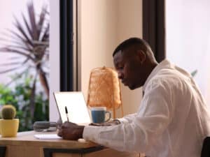 Young black man in white shirt writing memos on paper while sitting at table with netbook and doing freelance work