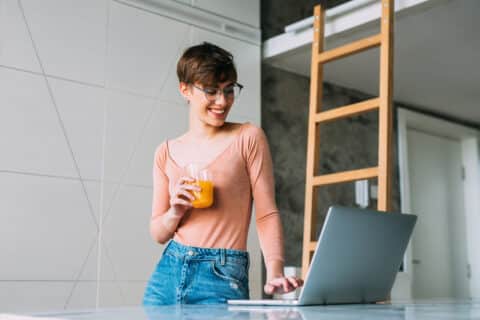 Beautiful young short-haired woman standing at her kitchen and looking at laptop.