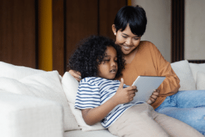 Beautiful smiling mixed race woman and her cute son sitting on couch and using tablet.